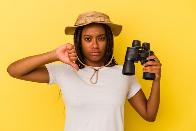 Kobieta młody odkrywca african american, trzymając lornetkę na białym tle na żółtym tle pokazując gest niechęci, kciuk w dół. Koncepcja niezgody.