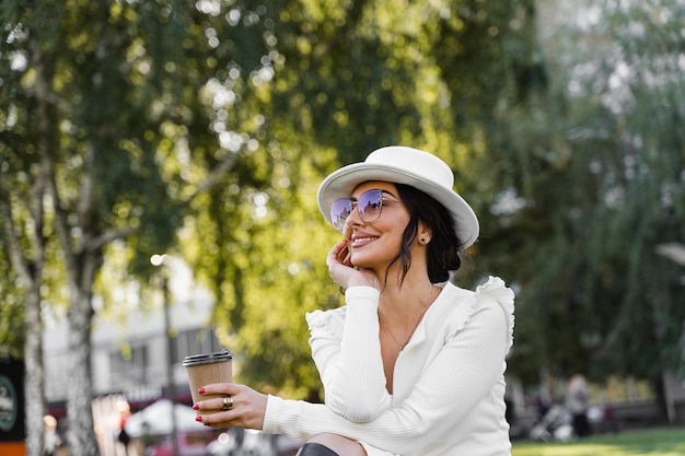 Kobieta moda cieszyć się kawą. Atrakcyjny model miał na sobie białą sukienkę, kapelusz i okulary przeciwsłoneczne, pozowanie na ulicy.