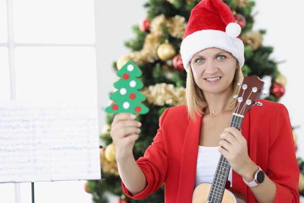 Kobieta muzyk w santa hat trzyma zabawkową choinkę i akompaniament muzyczny ukulele dla