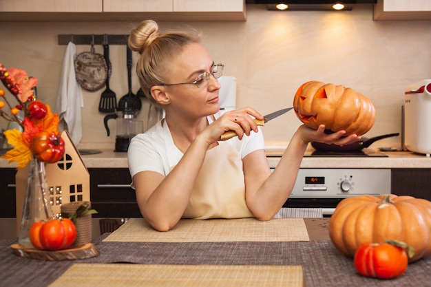 Kobieta Na Kuchni Rzeźbi Dynię Na Halloween W Pokoju Z Jesiennym Wystrojem I Lampą Domową. Przytulny Dom I Przygotowania Do Halloween.