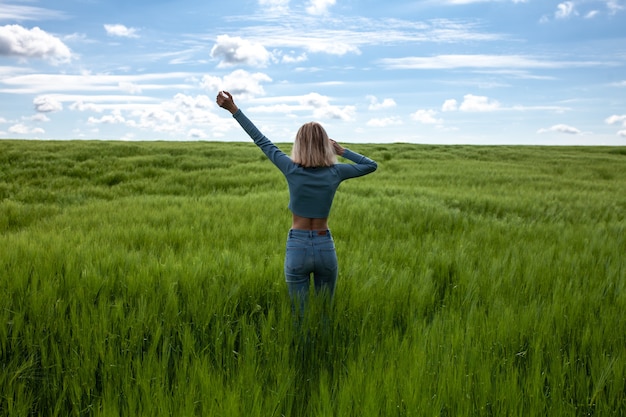 Kobieta na plecach z wyciągniętymi ramionami Szczupła młoda kobieta na środku pola pszenicy Jasne pole zielonej trawy Błękitne niebo z chmurami