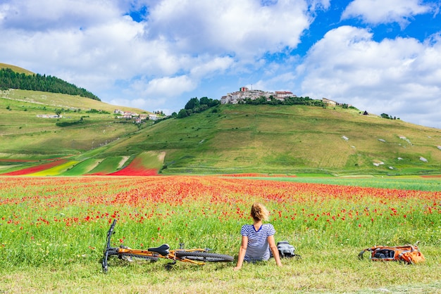 Kobieta na rowerze mtb w kwitnących polach uprawnych wyżyny Castelluccio di Norcia, słynnej kolorowej równiny kwitnącej w Apeninach we Włoszech. Uprawa roślin uprawnych i maków czerwonych.