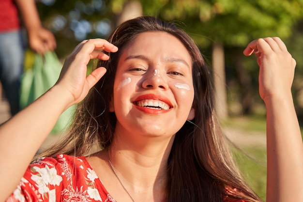Kobieta nakładająca krem przeciwsłoneczny na twarz Uśmiechnięta młoda Latynoska z długimi włosami i kremem przeciwsłonecznym na twarzy