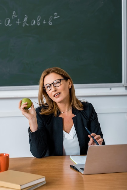 Zdjęcie kobieta nauczycielka w okularach siedząca przy biurku komputerowym i trzymająca jabłko w klasie