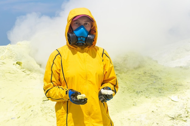 Kobieta naukowiec zajmująca się wulkanem z próbkami minerałów w dłoniach i respiratorze na tle dymiącego fumarolu na zboczu wulkanu