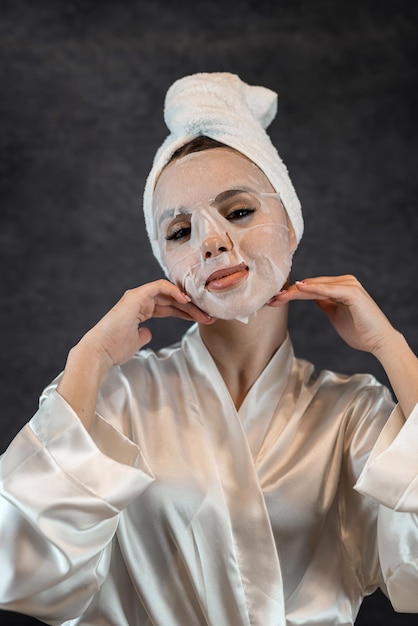 kobieta nosi biały ręcznik, nakładając maskę kosmetyczną w płachcie tkaniny do czystej pielęgnacji skóry twarzy odizolowanej na czarno Procedura koncepcyjna pielęgnacji skóry dla skóry w celu odmłodzenia i wyeliminowania defektów
