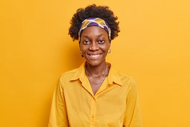 kobieta o ciemnej skórze uśmiecha się pozytywnie zadowolona z otrzymania komplementu wyraża wesołe emocje przychodzi na nieformalne spotkanie nosi casualową koszulę odizolowaną na żółtej ścianie