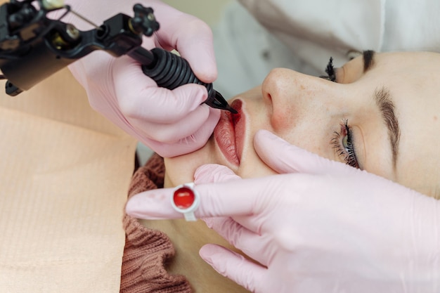 Kobieta o makijażu permanentnym na ustach w gabinecie kosmetycznym. Kosmetolog w rękawiczkach stosując tatuaż z maszyną, widok makro