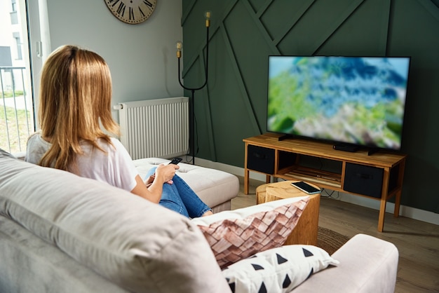 Kobieta odpoczywa w domu. Osoba ogląda telewizor na kanapie i przełącza kanały za pomocą pilota