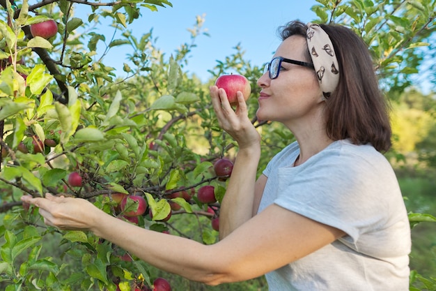 Kobieta ogrodnik ze świeżo zebranym czerwonym jabłkiem w ręku, tło jest drzewo z jabłkami. Samica zjada naturalne, przyjazne dla środowiska jabłko wyhodowane w przydomowym ogródku, kopia przestrzeń