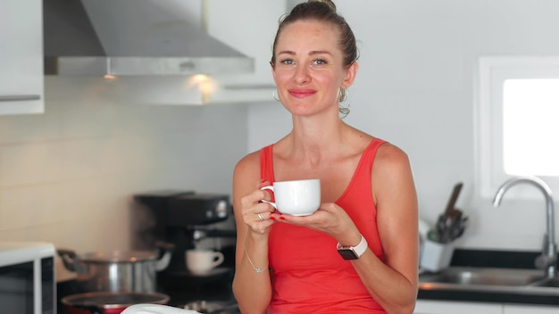 Kobieta pijąca herbatę z kubka w kuchni podczas gotowania posiłku i patrząca w kamerę
