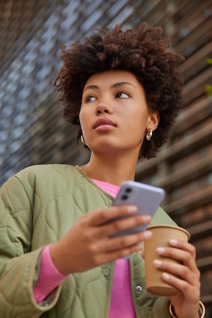 kobieta pije aromatyczny napój trzyma telefon komórkowy do rozmów online ubrana w zwykłe ubrania spacery na zewnątrz wraca do domu po studiach lub pracy