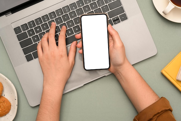 Kobieta pisząca na laptopie notebooka i trzymająca makietę białego ekranu smartfona nad biurkiem