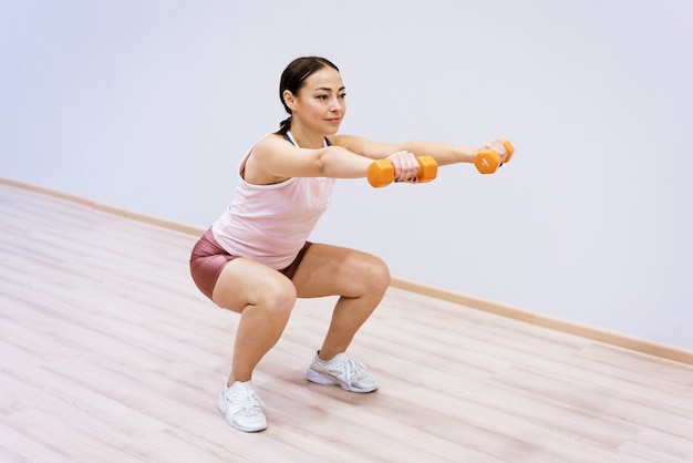 Kobieta pochodzenia kaukaskiego w sportowych przysiadach z hantlami w dłoni na tle...