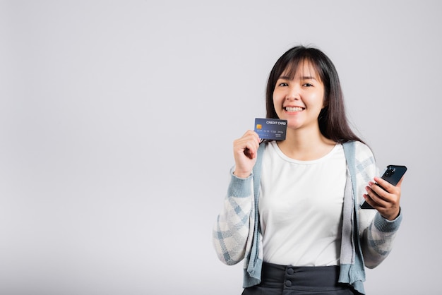 Kobieta podekscytowana uśmiechnięta trzyma telefon komórkowy i plastikową kartę kredytową debetową do płatności studio strzał na białym tle, szczęśliwa młoda kobieta korzystająca ze smartfona kupuj i płać zakupy online