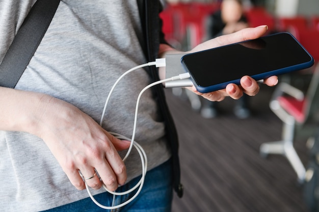 Kobieta podróżnik w swobodnym stroju ładuje baterię telefonu komórkowego za pomocą białego kabla USB z przenośnego urządzenia ładującego na lotnisku z bliska