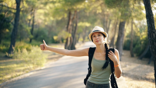 Kobieta podróżująca autostopem po słonecznej leśnej drodze Turystyczna dziewczyna szukająca przejażdżki, aby rozpocząć swoją podróż