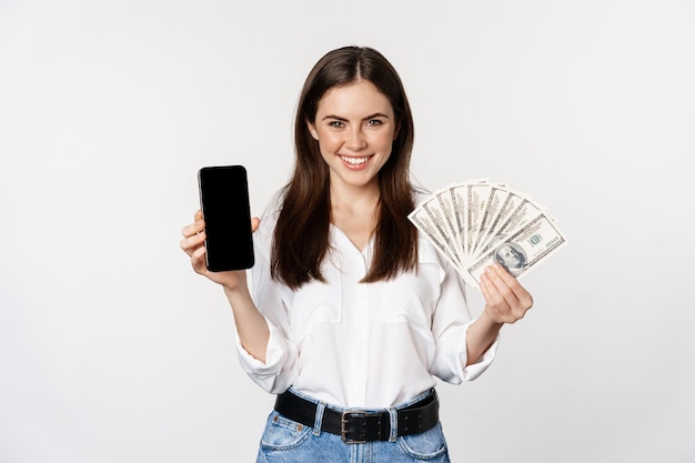 Kobieta pokazuje ekran telefonu komórkowego i gotówki, pieniędzy, koncepcji mikrokredytów i pożyczek bankowych, stojąc na białym tle.