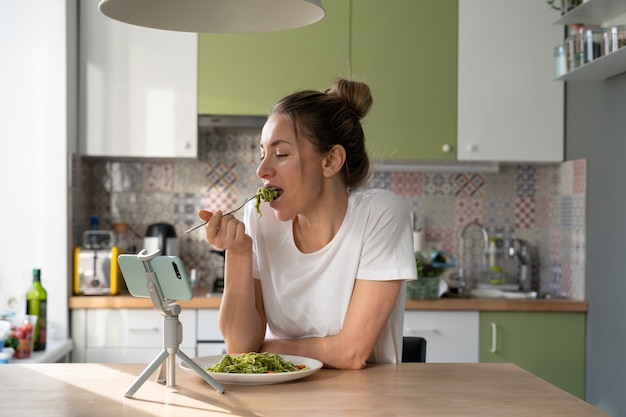 Kobieta powoli je makaron z wegetariańskim sosem pesto, oglądając film na smartfonie na statywie w domowej kuchni.