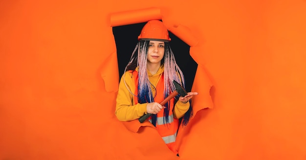 Kobieta pracownik budowlany z młotkiem wystającym z dziury na pomarańczowym tle Młoda kobieta w kombinezonie uderza narzędzie z ręki i patrząc na kamery