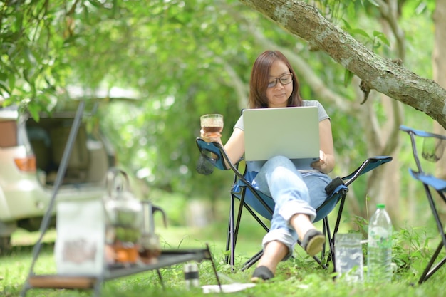 Kobieta pracująca z laptopem podczas biwakowania, w okularach i szarej koszulce, trzymająca filiżankę kawy na krześle kempingowym, patrząca na laptopa na nogach, podróżująca i pracująca.