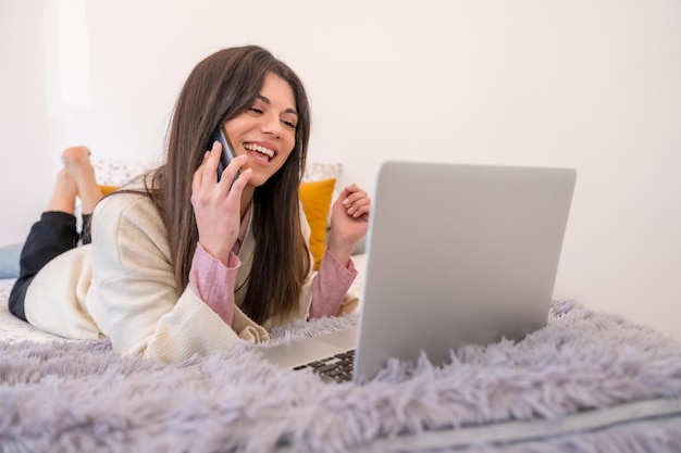 Kobieta pracująca z laptopem w łóżku telepraca telepraca uśmiecha się przez telefon