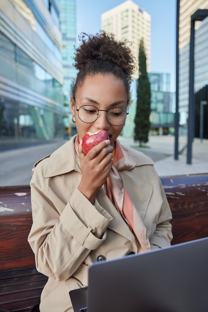 kobieta pracuje jako wolny strzelec na laptopie czyta artykuł online skupiony na ekranie zjada jabłko nosi okrągłe okulary do korekcji wzroku siedzi na ławce