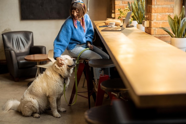 Kobieta pracuje na laptopie siedząc z psem w nowoczesnej kawiarni