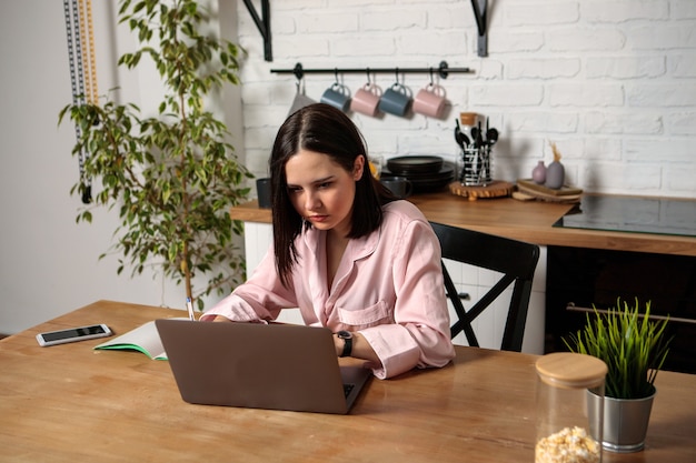 Kobieta pracuje w biurze zdalnie z domu w kuchni, siedząc przy komputerze. Kształcenie na odległość, edukacja i praca online. Studentka przygotowuje się do egzaminów.