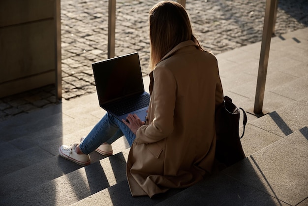 Kobieta pracuje zdalnie na ulicy miasta, korzysta z laptopa i siedzi na schodach