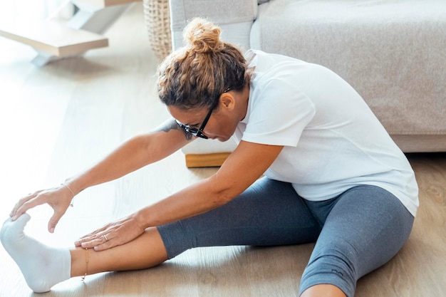 Kobieta praktykuje jogę lub pilates w swoim mieszkaniu, siedząc na podłodze w zwykłym ubraniu sportowym, koncentrując się na rutynie fizycznej, która promuje dobre samopoczucie, zdrowie i relaks umysłu