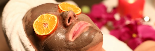 Kobieta przechodzi kurs odnowy biologicznej i oczyszczanie twarzy. Klientka leży na stole do masażu z włosami owiniętymi w ręcznik, maską węglową na twarzy i pomarańczowymi kręgami przed oczami.
