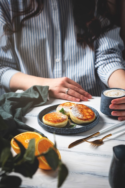 Zdjęcie kobieta przy stole z pięknym i smacznym śniadaniem. naleśniki z serem na talerzu, filiżance z kawą i pomarańczami na białym drewnianym tle.