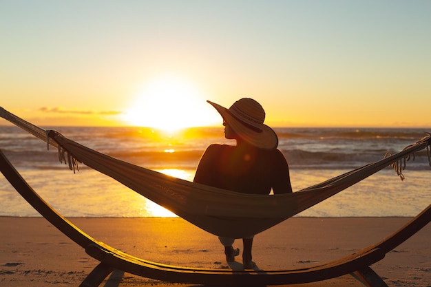 Kobieta rasy mieszanej na wakacjach na plaży siedzi w hamaku podczas zachodu słońca. zdrowy wypoczynek na świeżym powietrzu nad morzem.