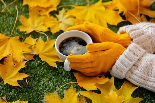 Kobieta ręce trzymać filiżankę kawy obok żółtych liści klonu na zielonej trawie