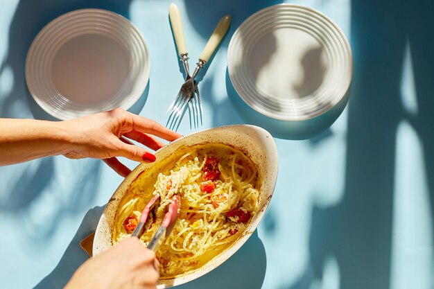 Kobieta ręka gotuje trendy wirusowe pieczone pomidory i feta z makaronem, fetapasta na niebieskim tle światło słoneczne, hardlight. Nowe, popularne danie obiadowe, widok z góry, miejsce na kopię.