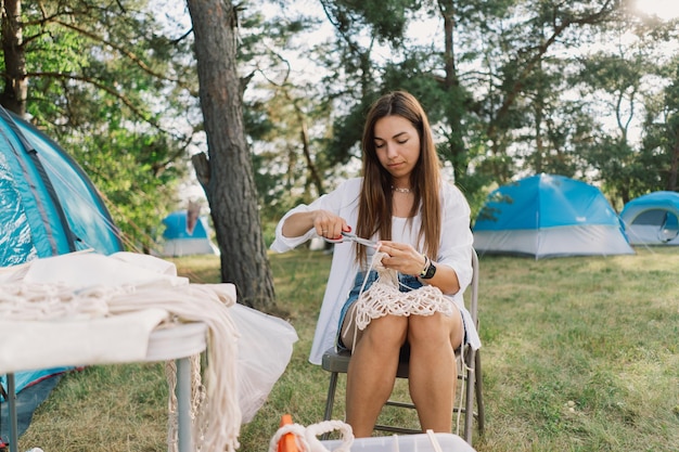 Kobieta robi na drutach torbę techniką makramy na świeżym powietrzu w pobliżu namiotów. Hobby na świeżym powietrzu. Tkanie makramy