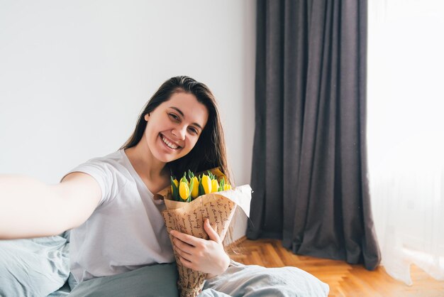 Kobieta robi sobie selfie z bukietem żółtych tulipanów w łóżku rano.