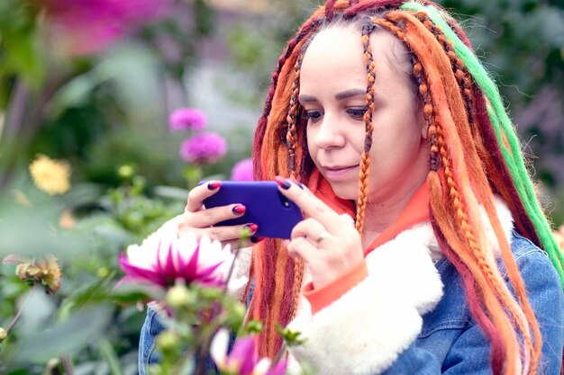 Kobieta robi zdjęcia na smartfonie w parku Pozytywna młoda kobieta hipster z długimi dredami w swobodnych ubraniach fotografująca kwiaty na telefonie komórkowym w parku