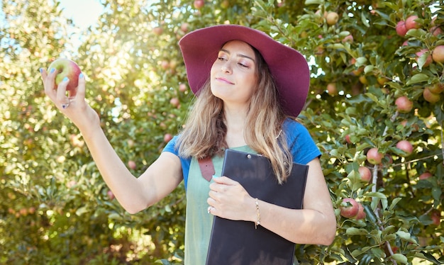 Kobieta rolnik sprawdzający produkty na farmie jabłek stojącej w sadzie w słoneczny dzień ze schowkiem do robienia notatek Młody pracownik gospodarstwa trzymającego owoce podczas żniw Rolnictwo ekologiczne
