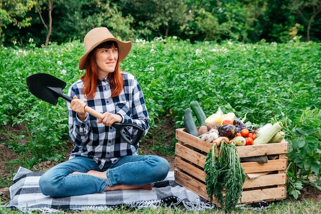 Kobieta rolnik z łopatą siedząca w pobliżu świeżych organicznych warzyw w drewnianym pudełku na tle veget