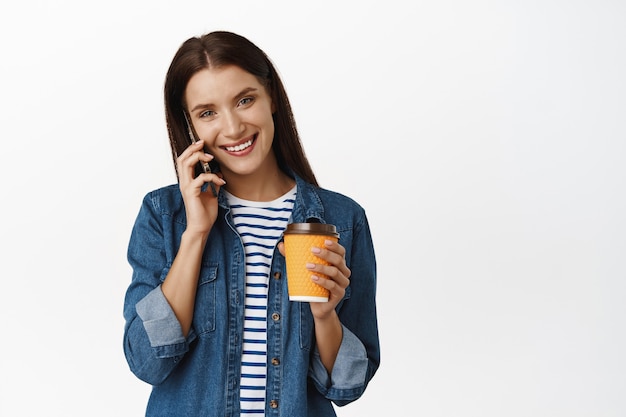 kobieta rozmowy telefonicznej i picia kawy na wynos. Ludzie biznesu. Uśmiechnięta kobieta używająca smartfona i trzymająca żółtą filiżankę z kawiarni na białym