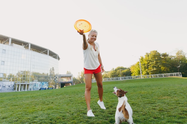 Kobieta rzucająca pomarańczowy latający dysk małemu zabawnemu psu, który łapie go na zielonej trawie. Mały Jack Russel Terrier zwierzak gra na świeżym powietrzu w parku. Pies i właściciel na świeżym powietrzu.