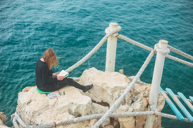 Kobieta siedząca na krawędzi i rysująca obraz mostu wiszącego z widokiem na morze