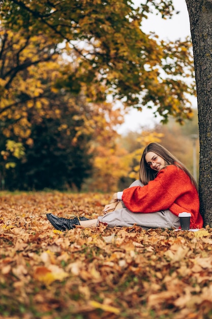 Kobieta siedząca pod drzewem na trawie pokrytej jesiennymi liśćmi i pijąca kawę