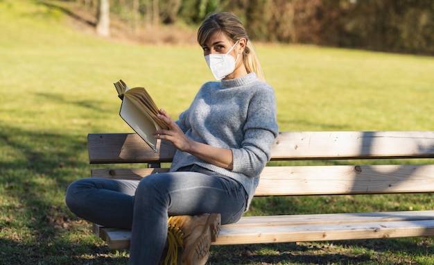 Kobieta siedzi na ławce w parku i czyta książkę z maską ochronną, aby zapobiec innym zakażeniom koronawirusem COVID19 i SARS cov 2 obraz koncepcji dystansu społecznego
