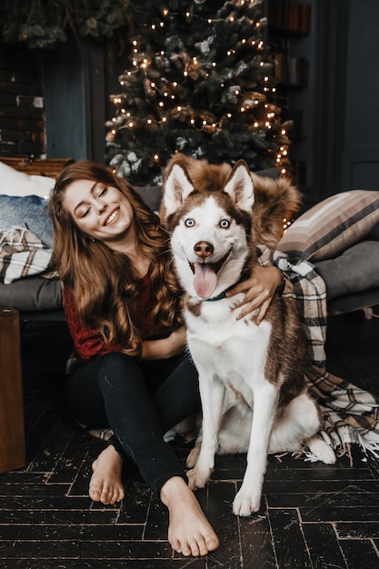 Kobieta siedzi na podłodze obok husky przy choince.