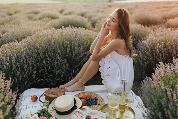 Kobieta siedzi na stole piknikowym na polu lawendy, otoczona kwiatami.