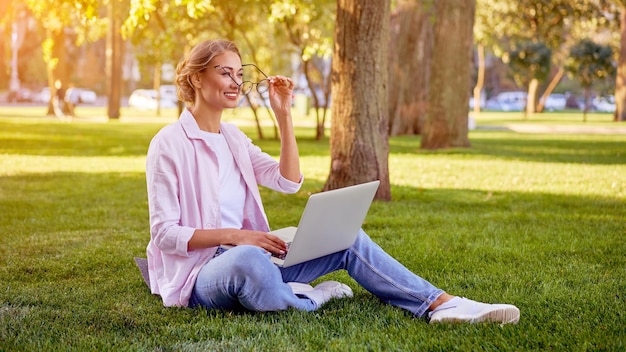 Kobieta siedzi na trawie w letnim parku za pomocą laptopa Biznesmen pracujący zdalnie na zewnątrz