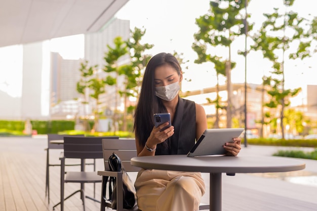 Kobieta siedzi na zewnątrz w kawiarni restauracja dystansu społecznego i nosi maskę na twarz, aby chronić przed covid 19 podczas korzystania z telefonu i tabletu cyfrowego poziomego strzału.